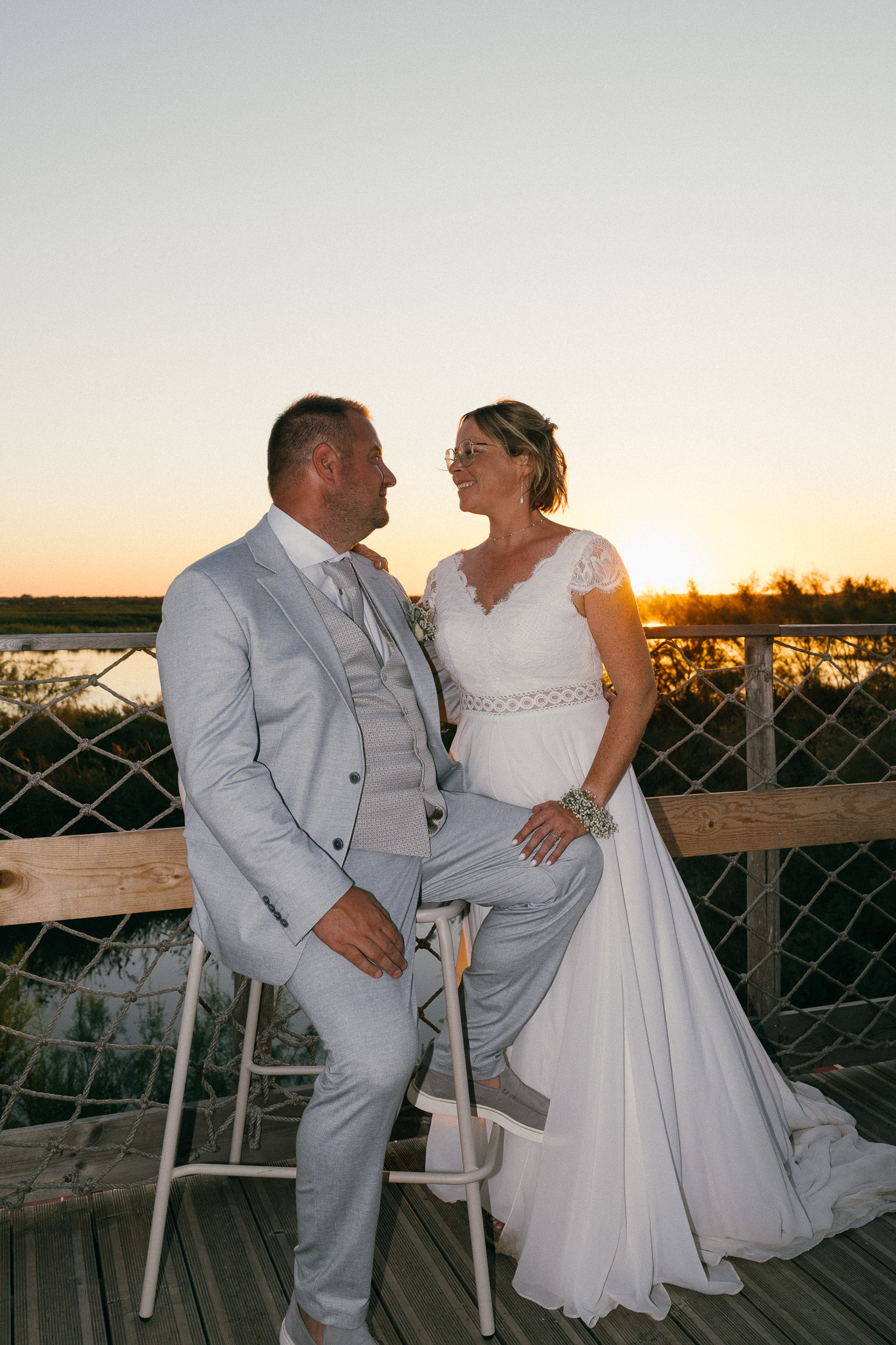 A full-length portrait of the newlyweds looking at one another by the water in evening light.