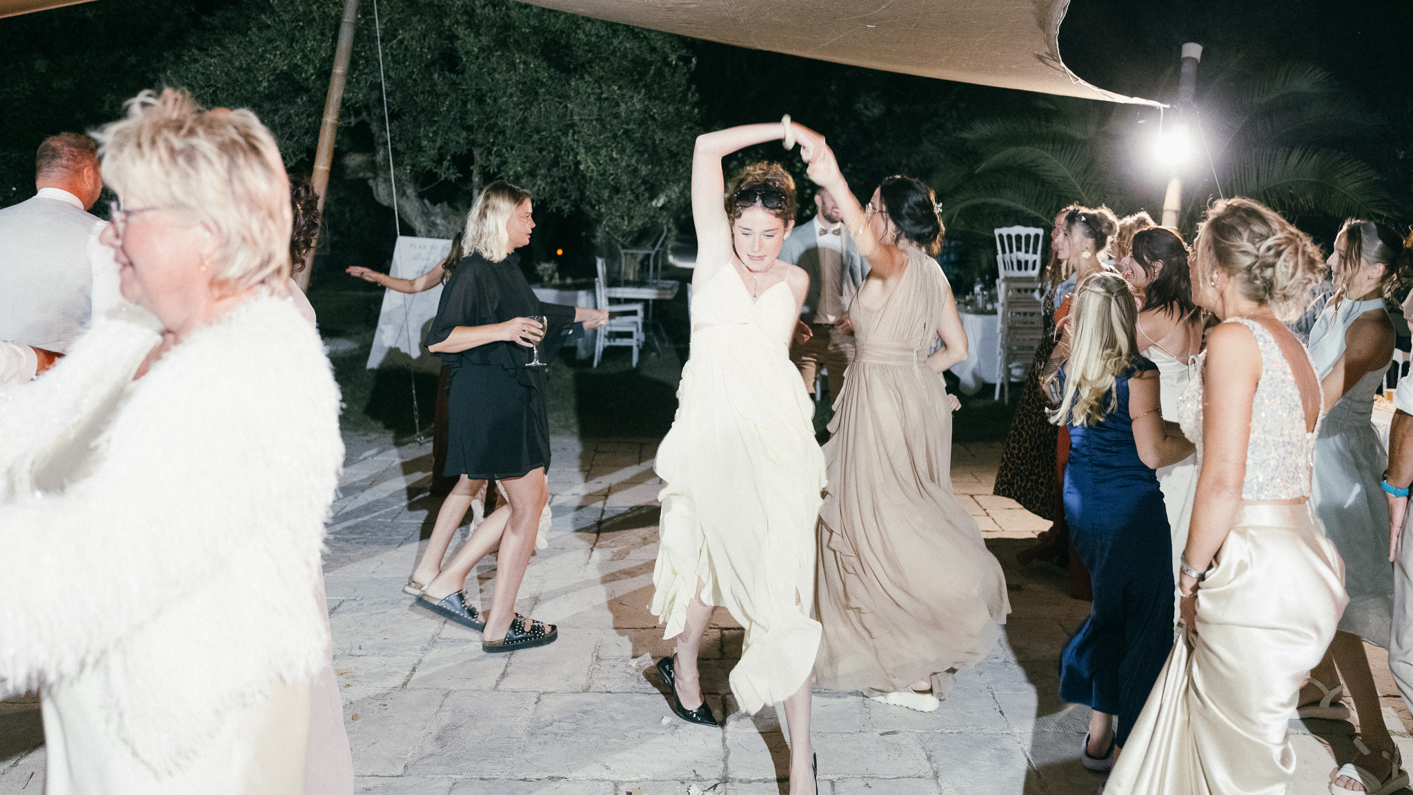 The bride dances with her arms raised, surrounded by guests at night.