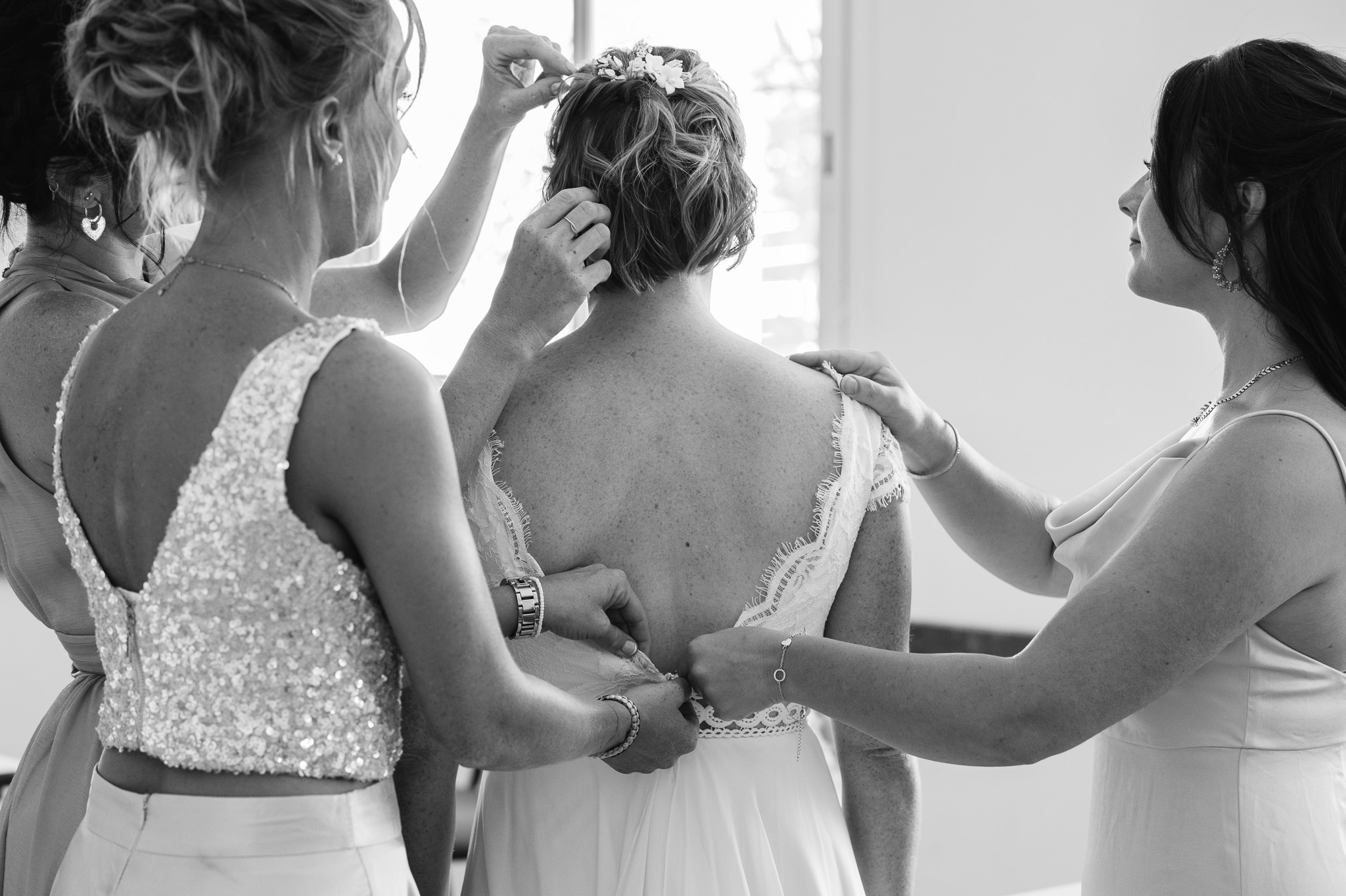 Two loved ones help the bride adjust her hair and dress, in black and white.