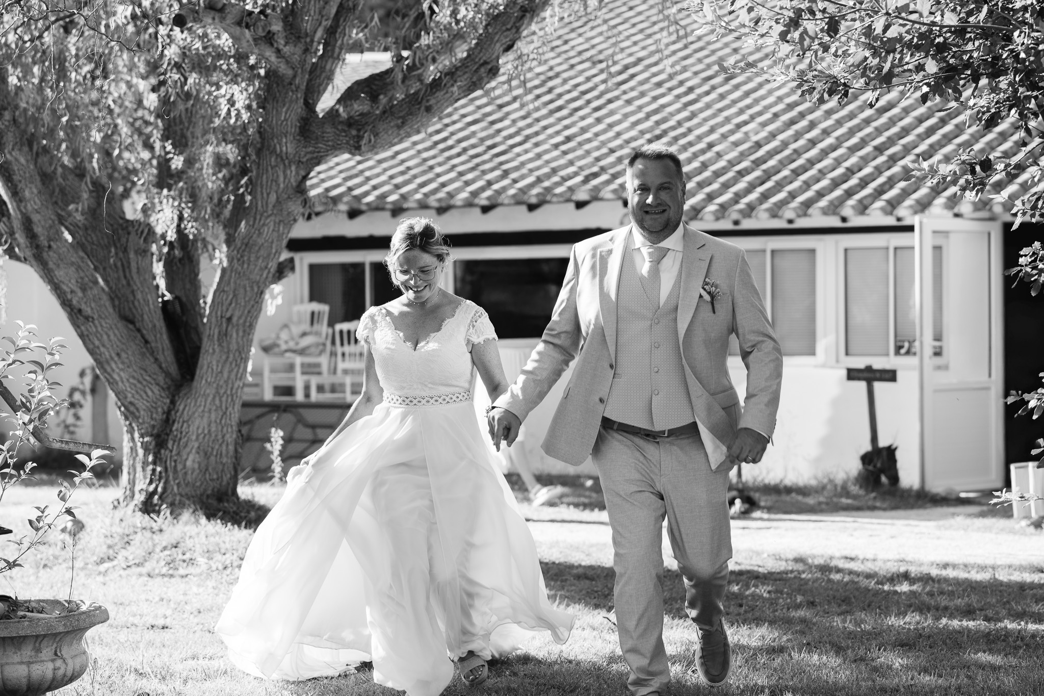 The newlyweds walk hand in hand beneath a tree, in black and white.