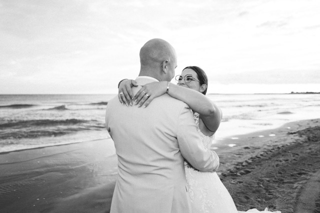 The bride looking at her partner as they embrace beside the sea, in black and white.