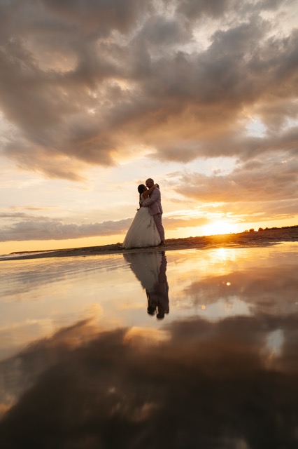The couple and their reflection in the water beneath a wide sunset sky.