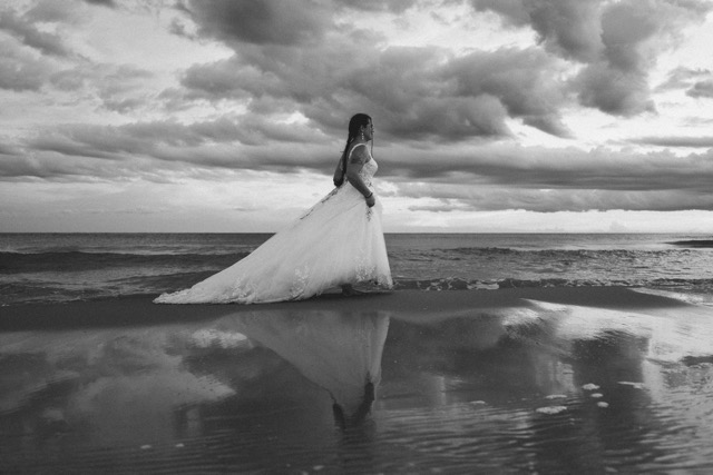 The bride’s silhouette and reflection by the sea beneath a cloudy sky, in black and white.