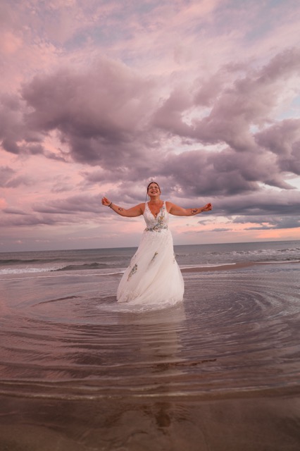 The bride with her arms open, her dress moving in the water at dusk.